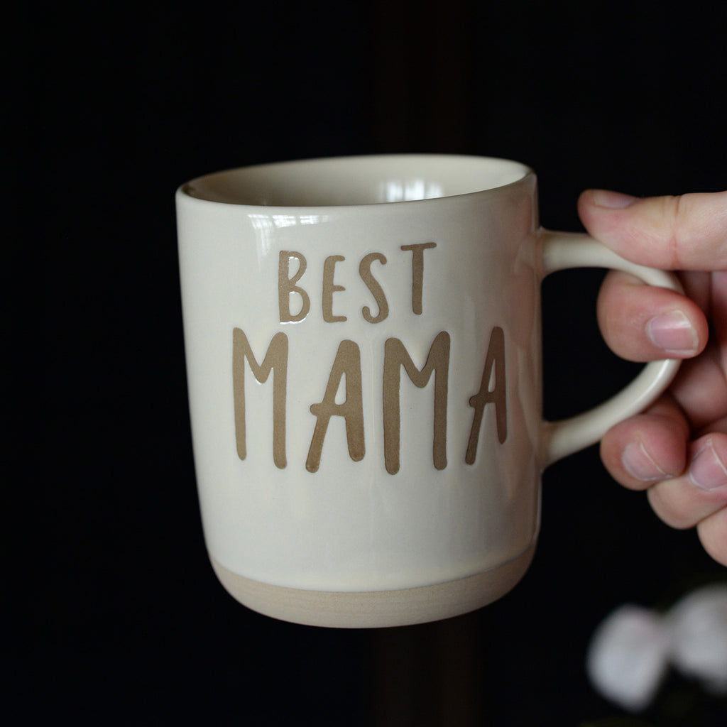 Beige mug with 'BEST MAMA' text held by a hand against a dark background