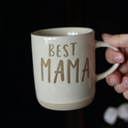 Beige mug with 'BEST MAMA' text held by a hand against a dark background