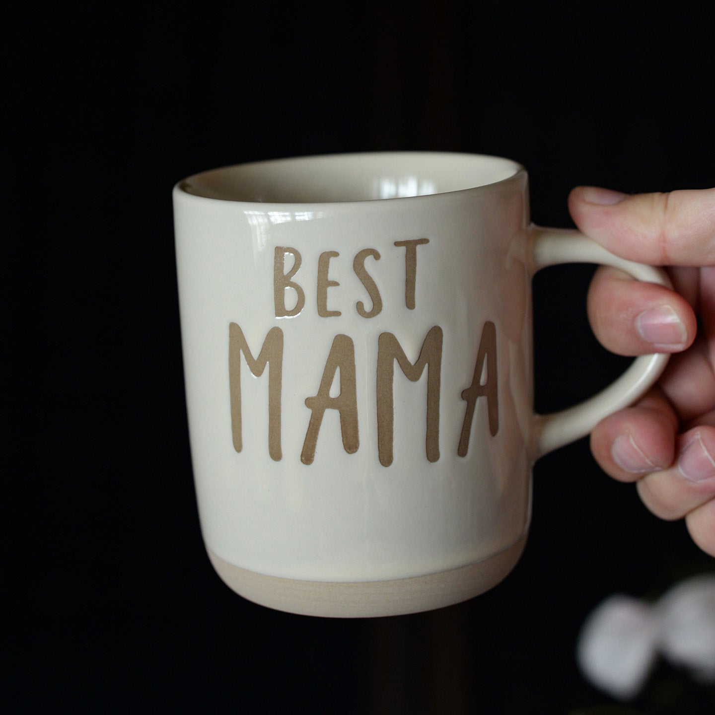 Beige mug with 'BEST MAMA' text held by a hand against a dark background