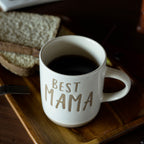 Beige mug with 'BEST MAMA' text on a wooden tray with toast and coffee.