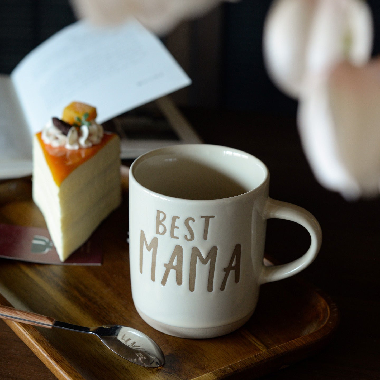 A beige mug with 'BEST MAMA' text on a wooden tray with a slice of cake and a spoon.